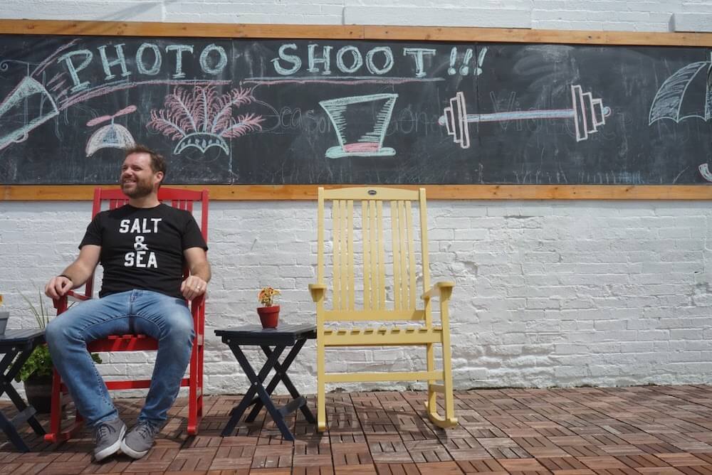 A man sits on a red chair against a chaolboard backgoround that says Photo Shoot