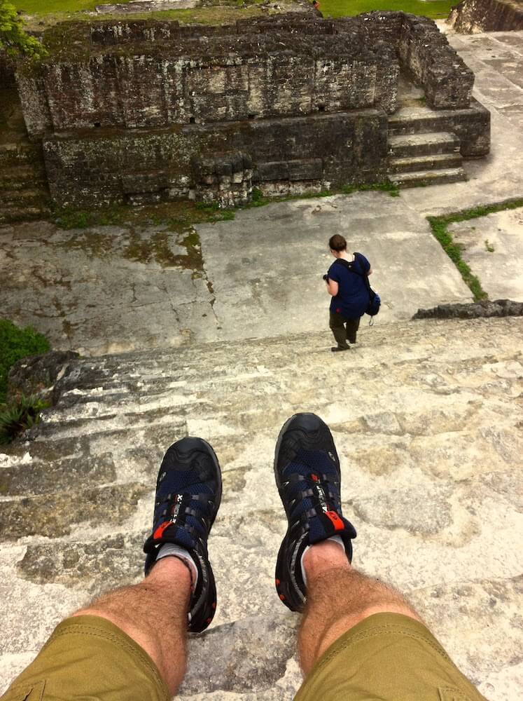 Men's feet are shown at the top of a pyramid in Tikal Guatemala looking down. A woman climbing down the pyramid can be seen at the bottom. 