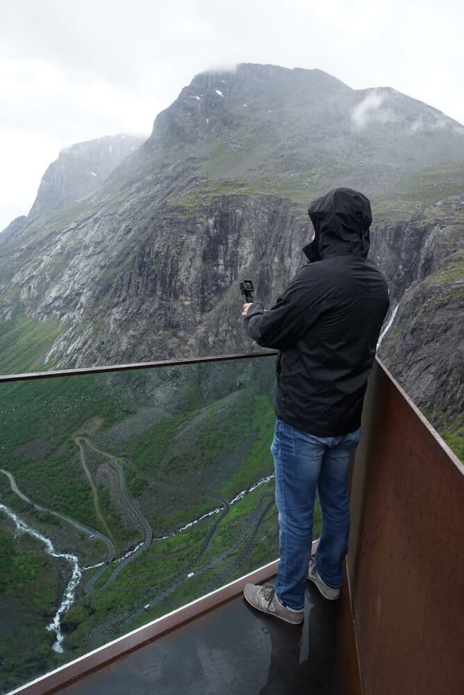 The back of a man holding a go pro overlooking the trollstigen on a viewing platform in Norway. The man is wearing a black rain jacket and jeans.