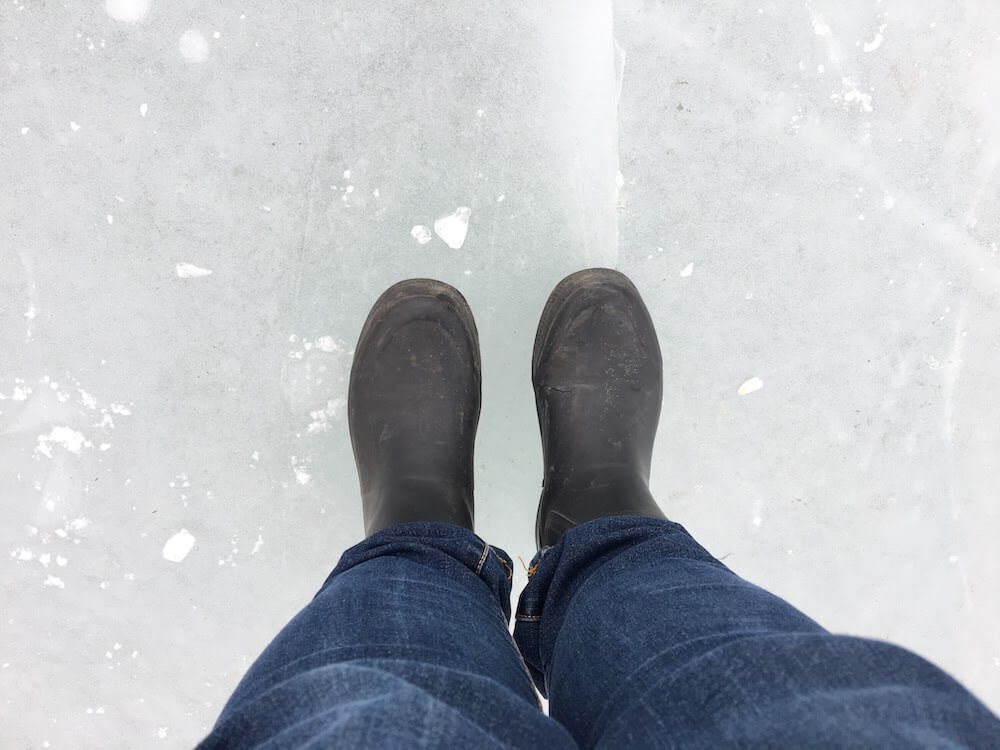 A pair of woman's legs and feet in jeans and black rubber rain boots standing on a frozen lake