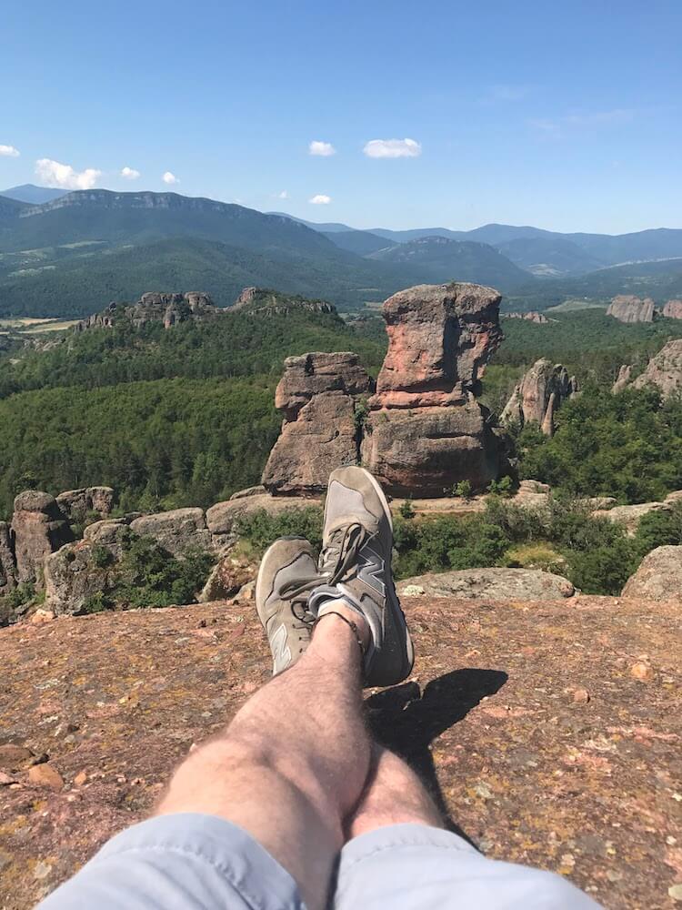 A man's legs wearing grey New Balance shoes can be seen at the top of Belogradchik Fortress in Bulgaria. Interesting rock formations are in the background.