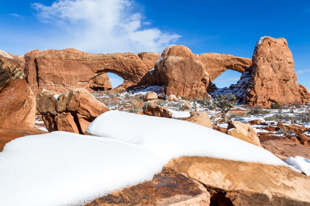 North and South Windows from Turret arch in Arches National Park, lit by sunlight and blue sky in the background with a bit of snow on the ground
