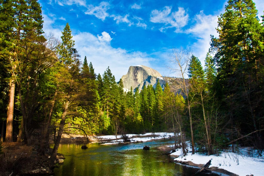 view of lake in sunshine with a bit of leftover snow in surrounding trees and mountains in Yosemite national park